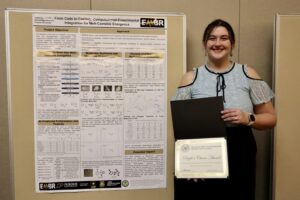 Person holding an award certificate standing next to an academic project poster.