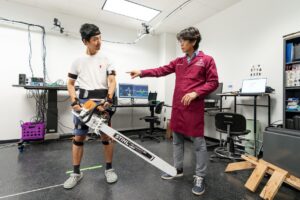 Man holding a long chainsaw and exoskeleton standing next to a man in a maroon lab coat inside of a laboratory setting.