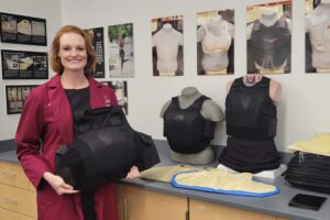 Dr. Porter standing in a room wearing a red lab coat, holding women's body Armor, with additional women's body armor on the countertop behind her. Several torso photos are on the wall above the countertop.