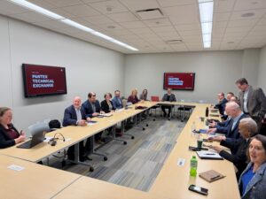 15 people in a room seated in a "horseshoe" with TV screens on the wall with "Pantex Technical Exchange" on the screens.