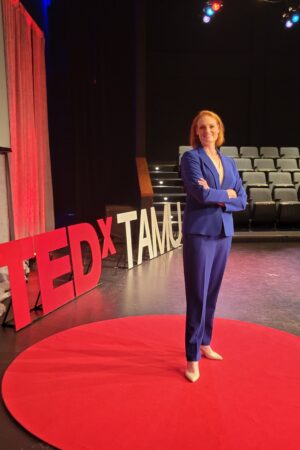 Woman in blue suit standing on a stage inside of a red circle, words "TEDxTAMU" in the Background.