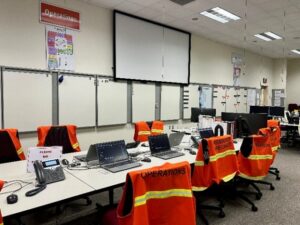 A room with a large white screen, the words "Operations" are on the wall, with several white boards underneath the sight. There is also a long set of tables with office chairs around it. Each chair has an orange safety vest on it, each vest has a title, some include "OPERATIONS," "RESOURCE SPECIALIST" among others that are unreadable. There are also telephones and computers on the table.