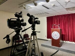 A podium with a maroon backdrop, two video cameras on tripods in the forefront.