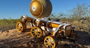 A six-wheeled robotic vehicle carrying a large ball on dirt.
